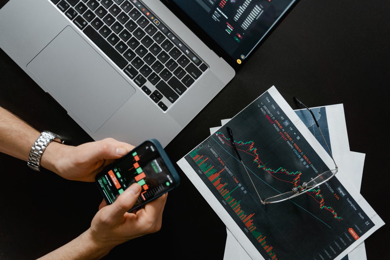 Des mains utilisent un smartphone à côté d’un ordinateur portable affichant des graphiques boursiers, illustrant le trading numérique.